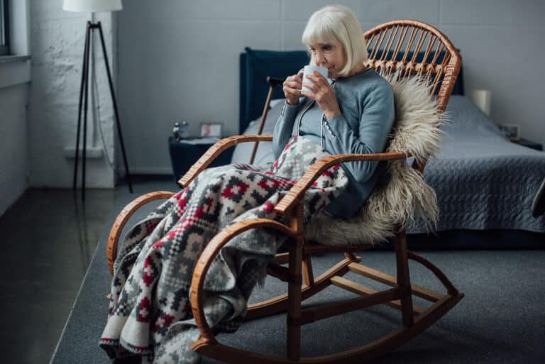 oudere vrouw die in rieten schommelstoel met deken zit en thuis koffie drinkt